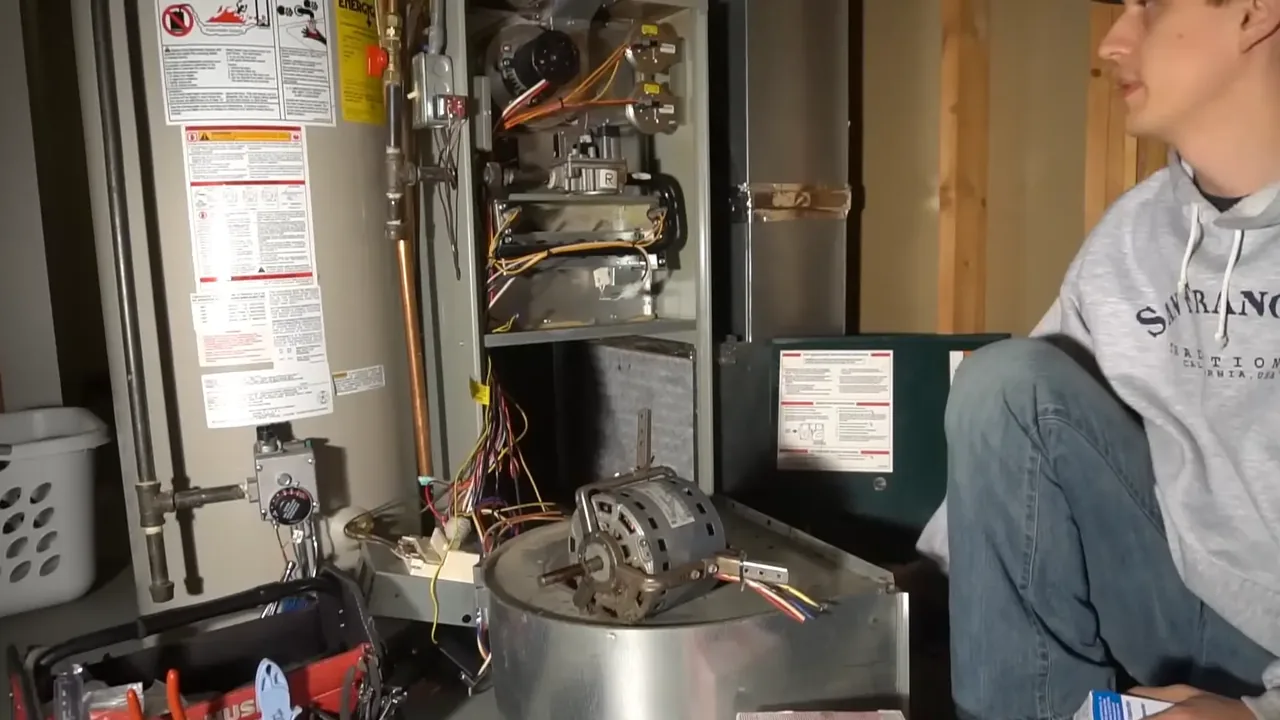 Furnace blower motor removed and resting on its housing beside an open furnace cabinet and a tool bag