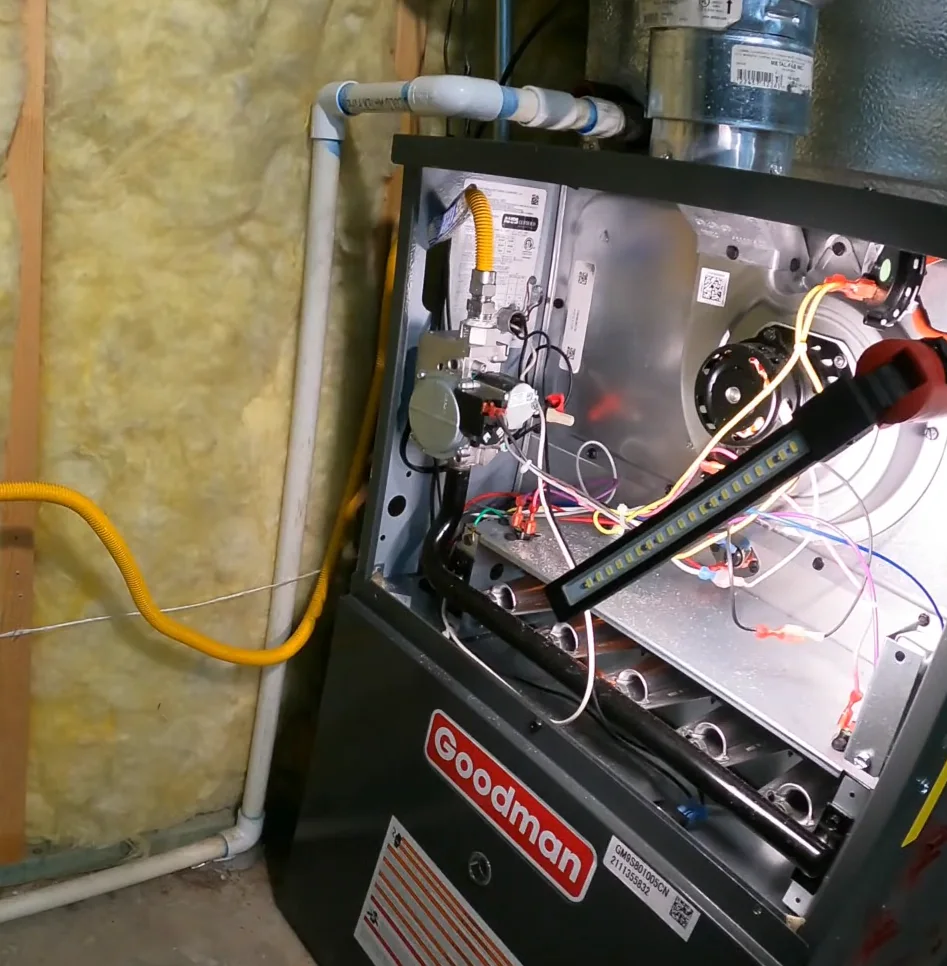 Open Goodman gas furnace with the front panel removed, showing the gas valve, yellow gas line, wiring and burner tubes lit by an LED work light