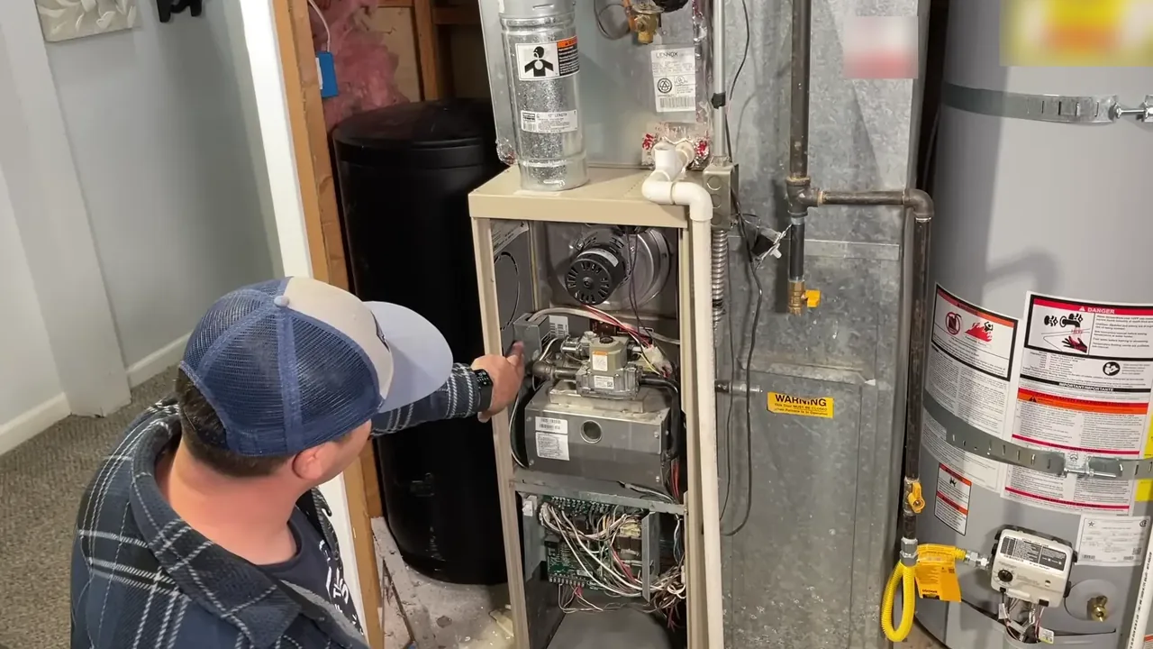 Technician in a baseball cap pointing into an open gas furnace near the gas valve, with a water heater beside it