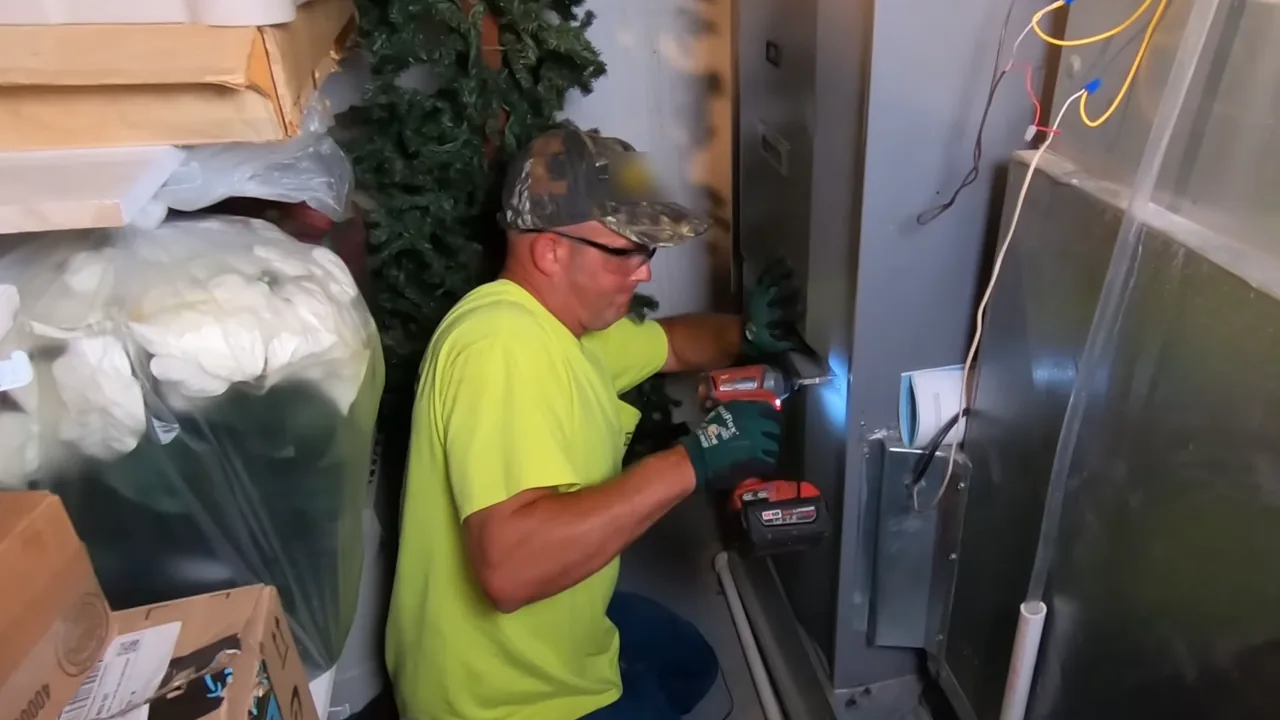 Technician in a high-visibility shirt and safety glasses using a cordless driver on the access panel of a furnace in a tight utility room